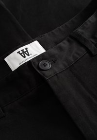 Pantalon en coton noir avec un design élégant, une fermeture à bouton et une étiquette portant l'inscription « DOUBLE V BY WOOD WOOD ». Détail du tissu texturé visible.