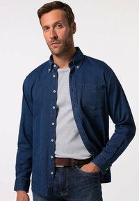 Camisa de denim en azul oscuro con diseño de botones, bolsillo en el pecho y mangas largas, superpuesta sobre una camisa gris. Tejido texturizado con costuras visibles.