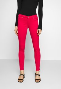 Jeans ajustados en denim rosa brillante, que cuentan con un cierre de botón y cremallera, bolsillos frontales y una silueta suave y entallada. Combinados con tacones negros.