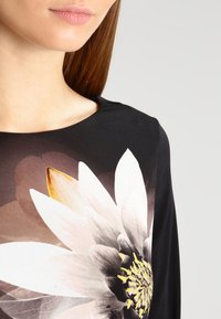 Czarny top z dużym białym nadrukiem lilii wodnej, w odcieniach przechodzących od ciemnego do jasnego, gładki materiał i elegancki dekolt.