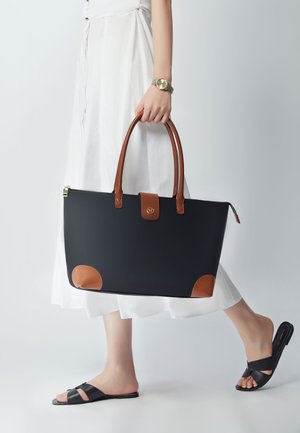 Black tote bag with brown leather accents, features a zip closure, gold hardware, and rounded corners. Model wears a white dress and sandals.