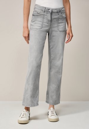 Vrouw in lichtgrijze cropped wide-leg jeans met voorzakken en witte sneakers tegen een neutrale beige achtergrond.