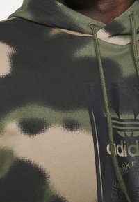 Huvtröja med kamouflagemönster i grönt och beige. Har ett strukturerat tyg och en svart logotyp tryckt på framsidan.