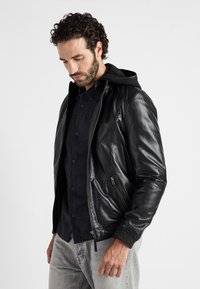 Schwarze Lederjacke mit Kapuze, Reißverschluss vorne, zwei seitlichen Taschen, gerippten Bündchen und einer glatten Oberfläche über einem dunklen Hemd und grauen Jeans.