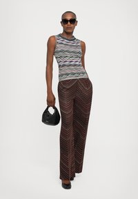 Kvinna i ärmlös flerfärgad randig topp, mönstrade bruna vida byxor, svarta klackar, solglasögon, hållande en liten svart handväska.