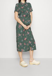 Abito floreale verde con maniche corte, vita con cintura e orlo al ginocchio, abbinato a zoccoli beige con suole in legno e dettagli metallici.
