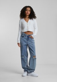 Gilet court blanc en peluche avec un col en V et une fermeture à bouton, associé à un Jean taille haute délavé bleu clair et des baskets blanches.