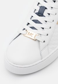 Baskets en cuir blanc avec des accents marine, arborant un logo doré MK sur un support de lacet, et des lacets plats blancs. Le panneau latéral texturé ajoute du détail.