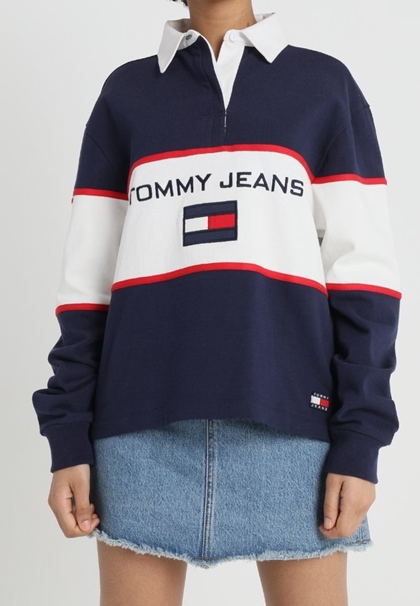 Maglietta in stile rugby blu navy e bianca con strisce rosse e logo "TOMMY JEANS" sul petto; abbinata a una gonna di jeans. Le maniche lunghe presentano polsini a coste.