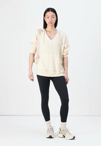 Helle beige, hüftlange Kapuzenjacke mit V-Ausschnitt, gerippter Textur und Fronttasche, kombiniert mit schwarzen Leggings und hellen Sneakers.