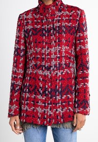 Donna che indossa un cappotto button-up testurizzato rosso, grigio e blu navy, con colletto alto e maniche lunghe, sopra dei jeans blu.