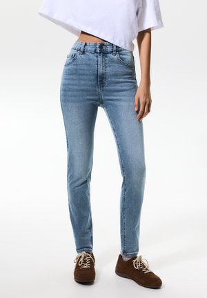 Persoon draagt lichtblauwe high-waist skinny jeans, een wit cropped top en bruine vetersneakers tegen een effen achtergrond.