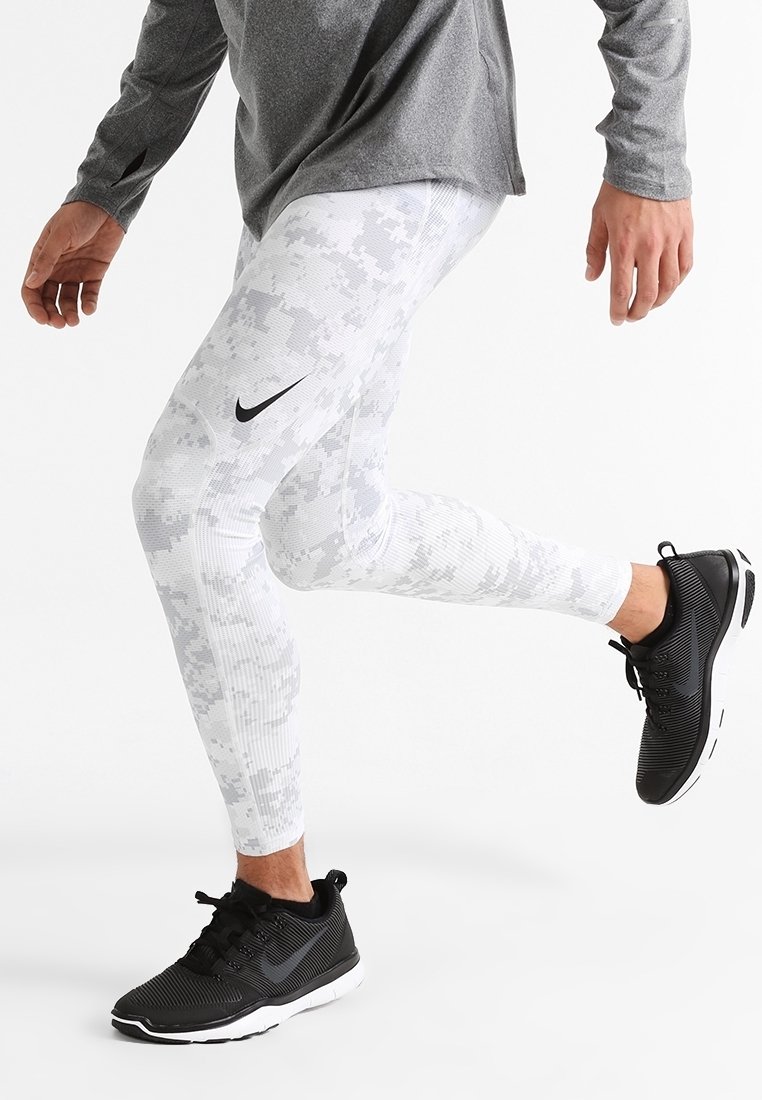 Leggings de compression blancs à motifs avec un logo Nike noir, associés à des chaussures de sport noires avec une tige texturée et une semelle blanche.