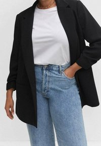 Henkilö, jolla on päällään musta bleiseri valkoisen t-paidan yllä ja vaaleansiniset korkeavyötäröiset farkut, toinen käsi taskussa yksinkertaista taustaa vasten.