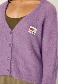 Paarse, fluffy cardigan met een v-hals en vijf knopen, over een olijfgroene top. Heeft een kleurrijke logo patch.