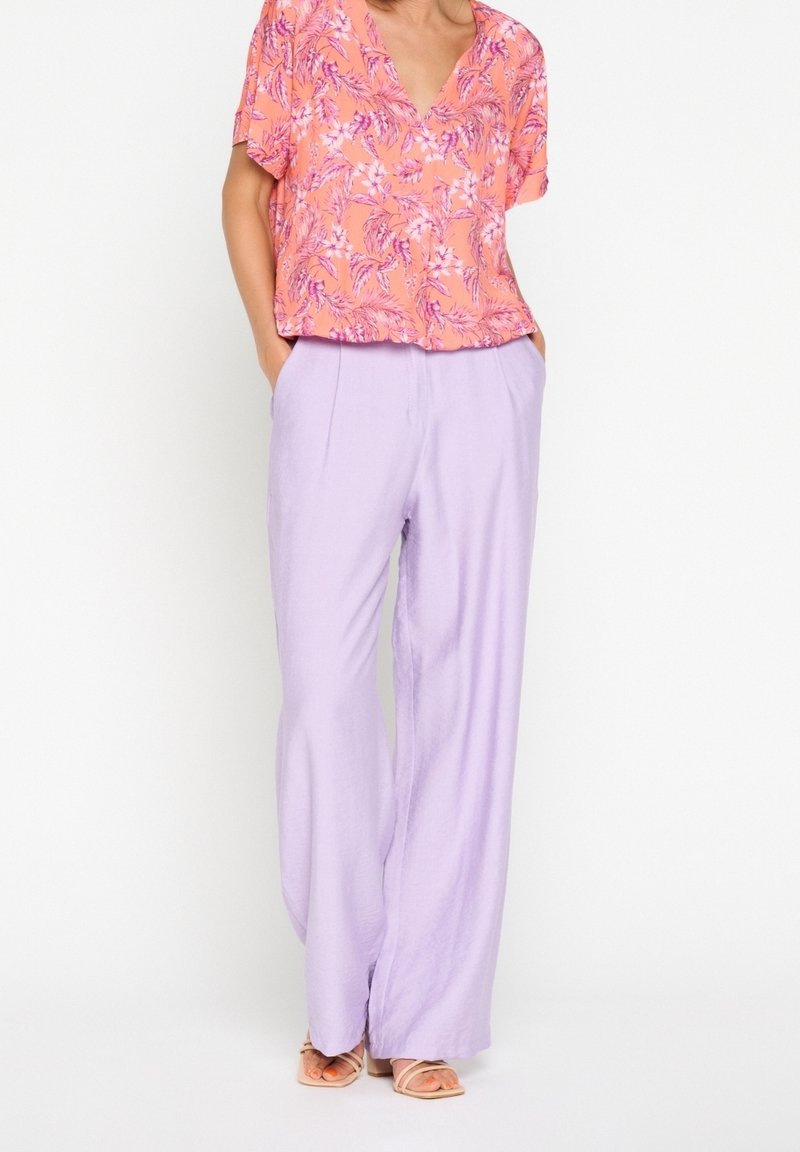Blusa a maniche corte di un luminoso corallo con motivo floreale, abbinata a pantaloni larghi in lavanda, realizzati in tessuto leggero. Sandali visibili.