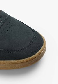 Zapatillas de ante negro con perforaciones para transpirabilidad, costuras azules contrastantes y una suela de goma marrón texturizada.