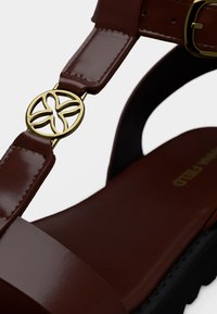 Καφέ δερμάτινο σανδάλι με φαρδιά λουρίδα, χρυσή στρογγυλή αγκράφα με λουλουδένιο σχέδιο, μαύρη υφασμάτινη σόλα, και λείο πέλμα.