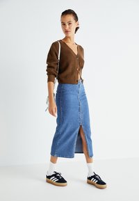 Rjav rebrast cardigan s popki, v kombinaciji z modrim denim midi krilom z razporkom spredaj. Celoten izgled dopolnjujejo črne teniske in bele nogavice.