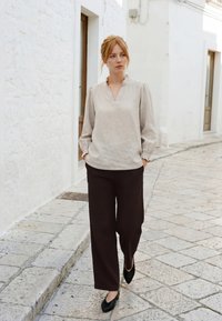 Femme aux cheveux roux attachés, portant un chemisier beige clair et un pantalon large foncé, marchant dans une rue étroite pavée de pierres avec des murs blancs.