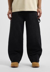 Pantalones de mezclilla negros holgados con una textura gruesa y un corte suelto, combinados con calzado beige que presenta una suela robusta y puntera redondeada.
