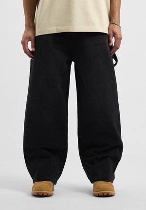 Pantaloni in denim nero oversize con una consistenza spessa e vestibilità ampia, abbinati a calzature beige con suola resistente e punta tonda.