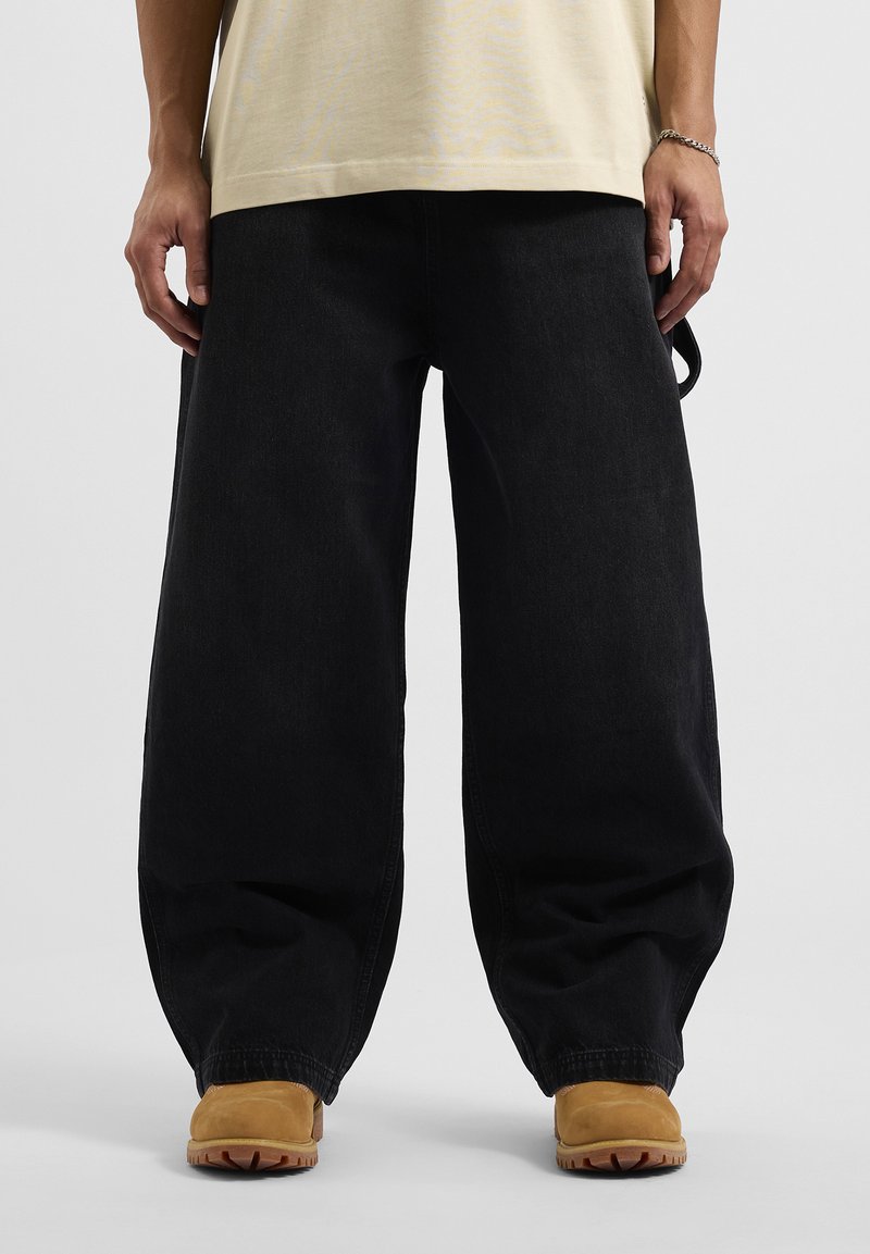 Pantalones de mezclilla negros holgados con una textura gruesa y un corte suelto, combinados con calzado beige que presenta una suela robusta y puntera redondeada.