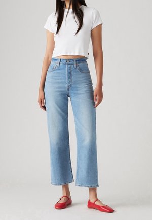 Vrouw draagt een wit cropped T-shirt, lichtblauwe high-waisted jeans met wijd uitlopende pijpen, en rode platte schoenen, staand tegen een effen achtergrond.