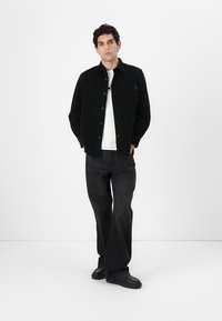 Chaqueta de pana negra con botones en la parte frontal, combinada con una camiseta blanca y jeans oscuros. El modelo lleva zapatillas negras y está de pie frente a un fondo liso.
