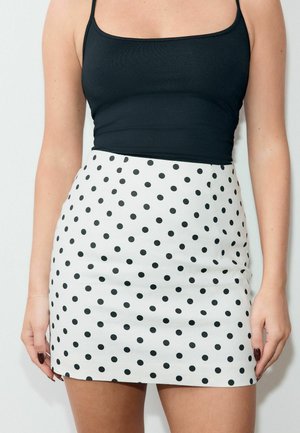 Žena v čiernom tielku bez rukávov a bielej mini sukni s čiernymi bodkami, stojaca na jednoduchom pozadí.