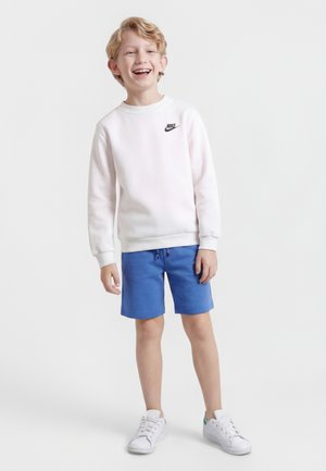 Ragazzo sorridente in piedi, che indossa una felpa bianca Nike, pantaloncini blu e scarpe da ginnastica bianche su uno sfondo grigio chiaro uniforme.