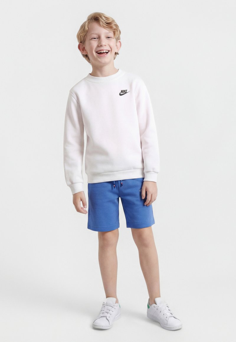 Χαμογελαστό αγόρι που στέκεται, φοράει άσπρο φούτερ Nike, μπλε σορτς και άσπρα αθλητικά σε απλό φόντο ανοιχτού γκρι χρώματος.