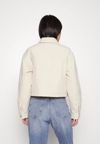 Cremefarbige, verkürzte Jacke mit Kragen, gestepptem Material und Knopfdetails. Der Träger kombiniert sie mit hellblauen Jeans.