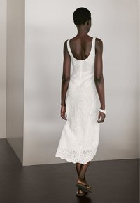 Witte jurk met een aansluitende corsage en een kanten, gegolfde zoom, aangevuld met eenvoudige sandalen en een dunne armband.