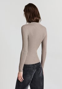 Person mit braunen Haaren, die ein enges, geripptes, beiges Langarmoberteil und lockere schwarze Jeans trägt, steht vor einem schlichten hellen Hintergrund.