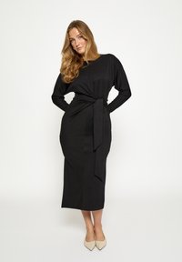 Mustast, pika varrukaga kleit, millel on vööga seotud detail ja mis on valmistatud siledast kangast. Disainil on sirge siluett ja põlveni ulatuv alumine serv.