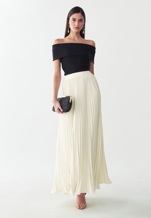 Donna con top nero a spalle scoperte e lunga gonna plissettata crema, tiene una pochette nera, in piedi davanti a uno sfondo semplice.