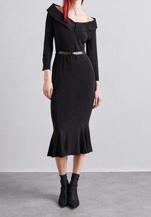 Kvinde iført en tætsiddende sort off-shoulder midi kjole med trekvartlange ærmer, sort bælte og sorte ankelstøvler med transparente sokker.