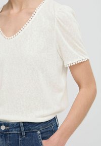 Haut blanc texturé à manches courtes avec un col légèrement dégagé, présentant une bordure décorative le long du col et des manches. Associé à un jean en denim bleu.