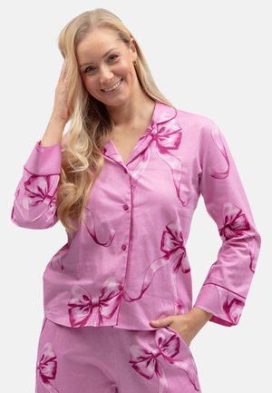Femme souriante aux longs cheveux blonds portant un pyjama rose décoré de grands nœuds rose foncé, une main touchant ses cheveux, l'autre dans la poche.