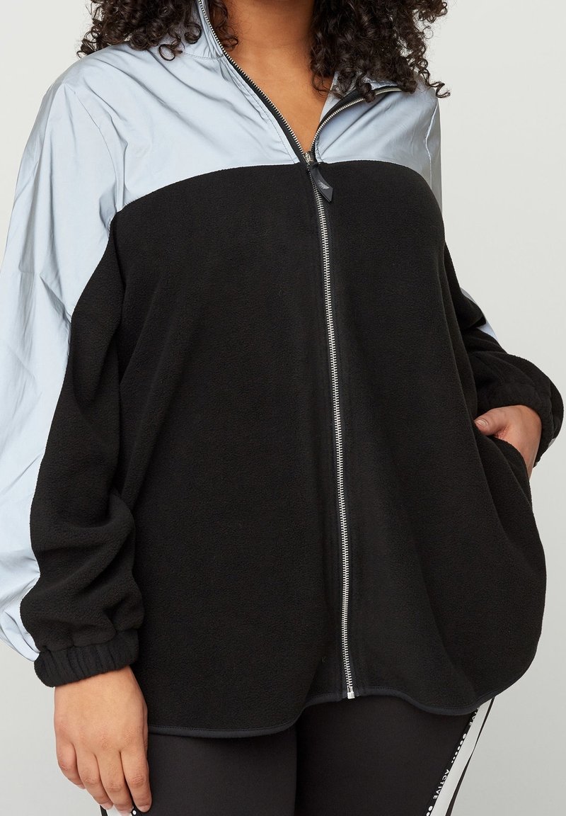 Veste en polaire noire avec partie supérieure en nylon bleu clair, fermeture éclair à l'avant et poignets côtelés. Offre une coupe décontractée et des poches latérales.