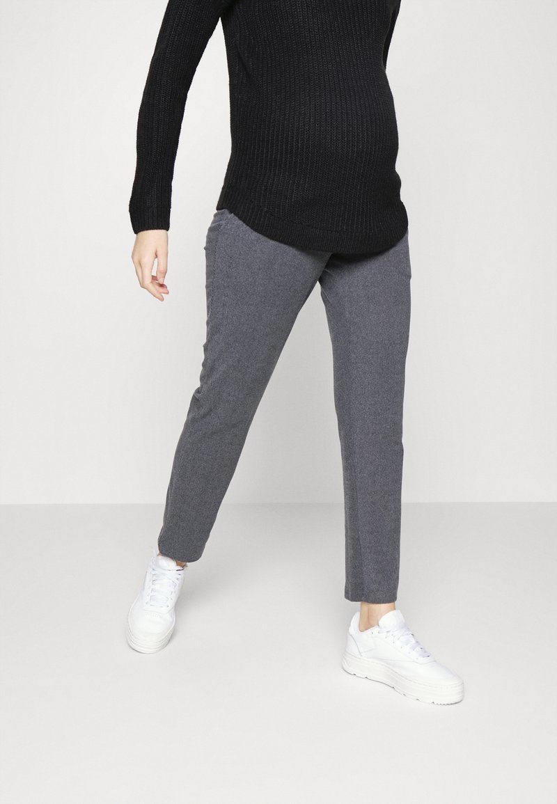 Personne portant un pantalon gris, des baskets blanches et un pull noir, debout les jambes écartées sur un fond blanc uni.