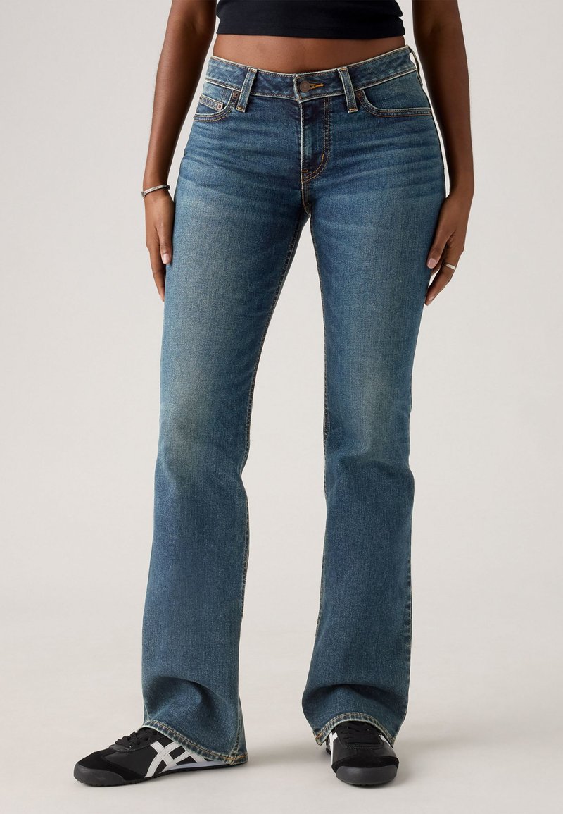 Kék harangszárú farmert viselő személy, fekete-fehér sportcipőben, fekete crop toppal, ezüst karkötővel és gyűrűkkel, amely egy sima háttér előtt áll.