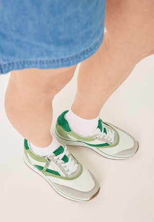 Baskets vertes et grises ensuédées et en mesh, avec des lacets et des semelles blanches. Assorties à des chaussettes blanches, visibles au-dessus d'un jean clair.