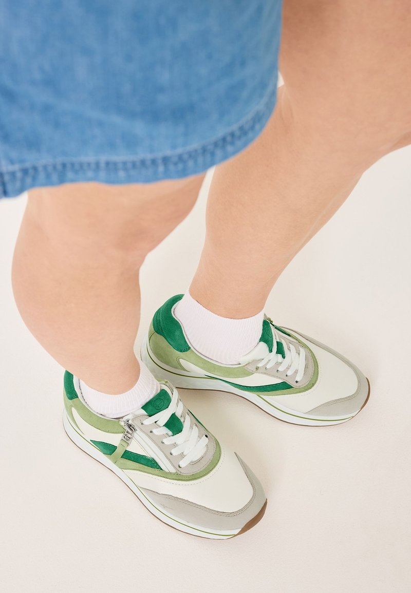 Sneaker verdi e grigie con materiali in suede e mesh, lacci e suole bianche. Combinati con calze bianche, visibili sopra un denim chiaro.
