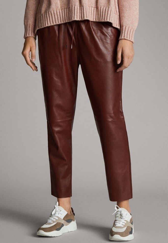 Personne portant un pantalon en cuir bordeaux, un pull beige rosé et des baskets blanches beiges, debout devant un fond gris uni.