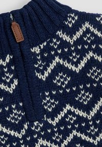 Marineblauer, gestrickter Pullover mit weißen geometrischen Mustern, hohem Rippenkragen und Reißverschluss, der mit einem braunen Lederakzent versehen ist.