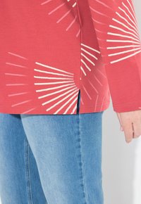 Chemise rouge à manches longues avec des motifs en forme de fan blanc, fentes latérales, associée à un jean bleu clair. Matière en coton, coupe décontractée, couture visible.