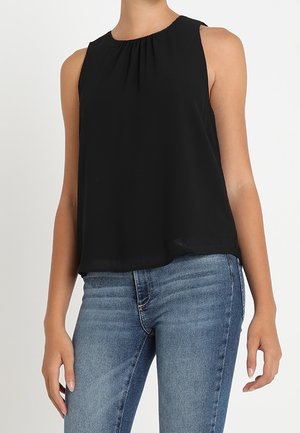 Ärmellose schwarze Bluse mit gerafftem Ausschnitt, kombiniert mit mittelblauen Jeans an einer stehenden Person von den Schultern bis zu den Hüften.