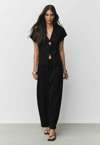 Schwarzes, kurzes Oberteil mit kurzen Ärmeln und Knopfdetail, kombiniert mit lockeren schwarzen Hosen und schwarzen Flip-Flops. Glatte Stofftextur.